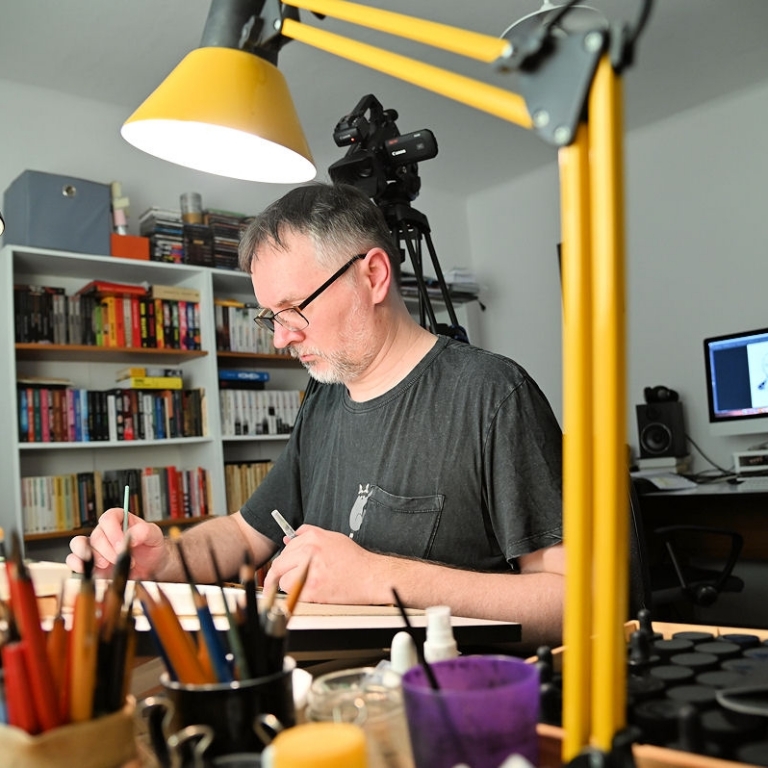 Maciej Mazur siedzi za biurkiem w czasie rysowania planszy komiksowej przed nim zółta lampka biurkowa