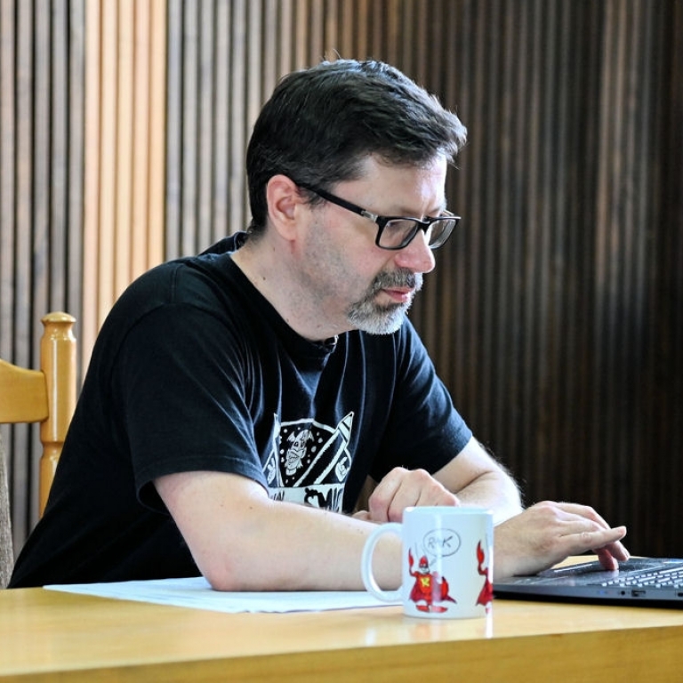 Jarosław Ejsymont siedzi przy stole przed laptopem przed nim stoi kubek Rzeszowskiej Akademii Komiksu